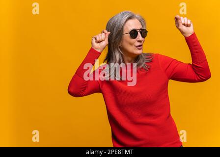 Donna gioiosa e dai capelli grigi in occhiali da sole che ballano e guardano lontano davanti alla macchina fotografica. Positiva donna matura di mezza età sorride, celebrando qualcosa di isolato su sfondo giallo Foto Stock