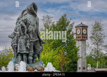 Fairbanks, Alaska, USA - 27 luglio 2011: Statua e fontana della prima famiglia sconosciute sul Golden Heart Plaza. Lato bambino e padre, con Torre dell'Orologio rotante Foto Stock