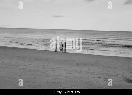 Tre visitatori si trovano sulla costa guardando la marea e le onde a Salisbury Beach, Massachusetts. L'immagine è stata acquisita in bianco e nero analogico Foto Stock
