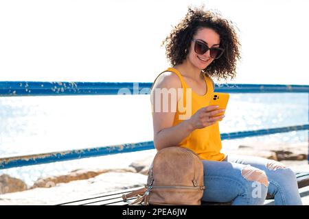 Giovane donna con capelli ricci positivi in abiti casual e occhiali da sole seduti sulla panca con zaino e sorridente mentre si naviga in smartphone contro il mare d Foto Stock