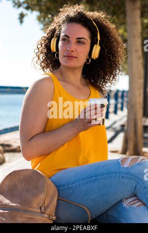 Donna giovane e pensosa dai capelli ricci in abiti casual e cuffie seduti sulla panca con zaino e caffè da asporto mentre riposa durante le vacanze estive Foto Stock