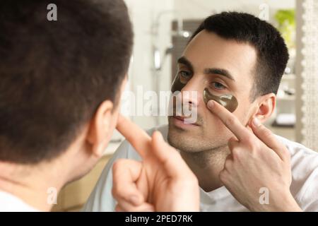 Giovane uomo che applica sotto gli occhi patch vicino a specchio a casa Foto Stock