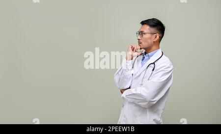 Medico maschile asiatico premuroso e professionale in abito bianco e occhiali, mano su mento, guardando da parte lo spazio vuoto, pensando a qualcuno Foto Stock