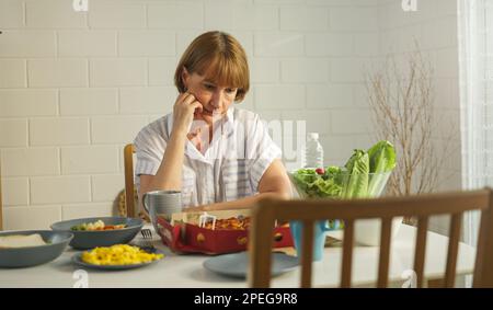 Nonna anziana in pensione rimanere a casa con il viso infelice seduto da solo sul tavolo da pranzo in casa. Depressa matura anziana donna rovesciato sensazione di lon Foto Stock