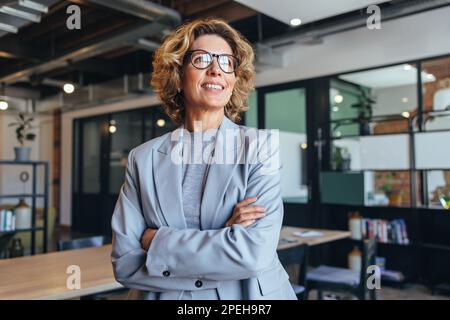 una donna d'affari con i suoi 40 anni guarda lontano con attenzione in un ufficio. Donna professionale in piedi con un vestito con le braccia incrociate. Foto Stock