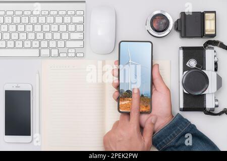 La mano di un uomo tiene un telefono su una scrivania piena di attrezzature, macchine fotografiche, telefoni e tastiere per computer. Foto Stock