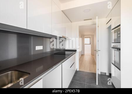 Moderna cucina high tech in bianco e nero con elettrodomestici e mobili moderni, con una porta aperta che si affaccia sul corridoio e una piccola camera con una finestra Foto Stock