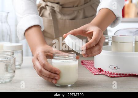 Donna con vaso di vetro di yogurt fresco gustoso a tavola di legno bianco in cucina, primo piano Foto Stock