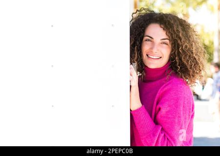 Felice femmina dai capelli ricci che indossa un maglione dolcevita rosa appoggiato sul muro mentre si trova sullo sfondo sfocato della strada cittadina nelle giornate di sole e lo Foto Stock