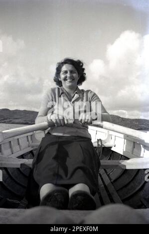 1950s, storica, fuori sul mare sulla costa, una giovane donna, in cima e gonna, in vacanza, divertendosi a remi su una barca di legno, Inghilterra, Regno Unito. Foto Stock