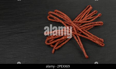 Salsiccia affumicata secca, bastoncini di salame sottile, Cabanossi, Kabanos, Embutido secco, Salsiccia di bratwurst sottile su fondo di pietra nera Foto Stock