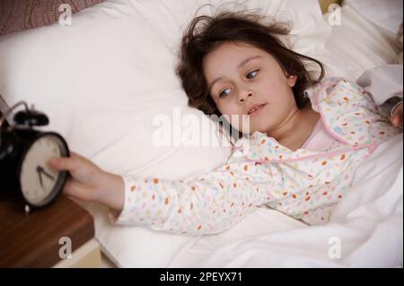 Adorabile bambina sdraiata in un comodo letto, svegliarsi e spegnere la sveglia la mattina presto Foto Stock