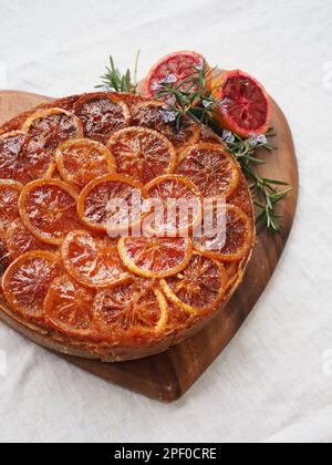 Primo piano di un'appetitosa torta rovesciata di arancia rossa su una tagliera a forma di cuore su una tovaglia di lino bianco Foto Stock