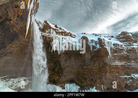 La cascata ghiacciata di Kvernufoss, con neve e stalagmiti bluastre con acqua che cade dalla vista come una grotta e ciottoli cielo nuvoloso in Islanda Foto Stock