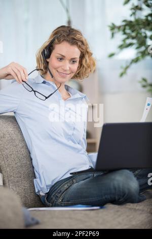 ragazza sorridente in cuffia sedersi sul divano guardando il portatile bere Foto Stock