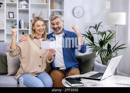 Coppia di famiglia felice, uomo e donna adulti maturi seduti a casa sul divano, celebrando la vittoria tenendo le mani in su soddisfatto con il budget della famiglia, dietro la famiglia di lavoro di carta con il portatile. Foto Stock