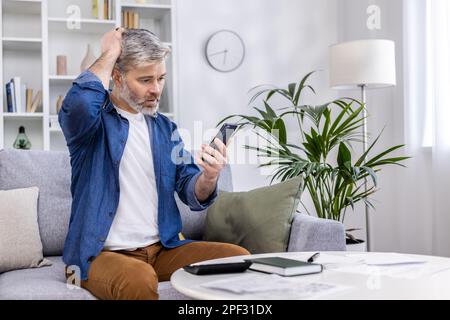 Uomo adulto anziano seduto sul divano da solo a casa, utilizzando lo smartphone nel soggiorno, leggendo le cattive notizie online dall'app al telefono, turbato e nervoso. Foto Stock