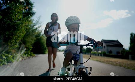 Madre insegnando al figlio di andare in bicicletta fuori in una strada urbana. Ragazzo di successo che indossa un casco di bilanciamento e pedalando da solo su una bicicletta. Han Foto Stock