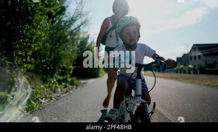 Madre insegnando al figlio di andare in bicicletta fuori in una strada urbana. Ragazzo di successo che indossa un casco di bilanciamento e pedalando da solo su una bicicletta. Han Foto Stock