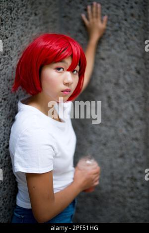 Teenage Girl in Cosplay Costume in piedi con piede e pendere su parete di cemento | Asiatico | profondità di campo | Wig Rosso Foto Stock