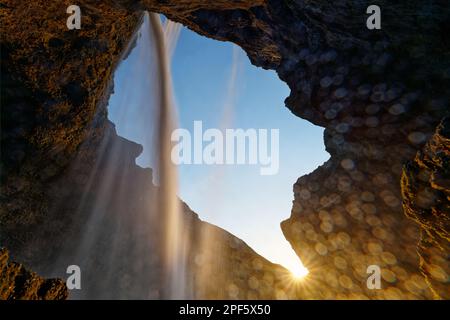 Vista diagonalmente dal basso attraverso una cascata circondata da rocce, il sole tramontante con un alone di raggi può essere visto al bordo dell'aperitivo panoramico Foto Stock