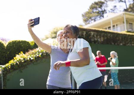 Felice diverse anziane amiche con racchette da tennis che prendono selfie al campo da tennis nelle giornate di sole Foto Stock