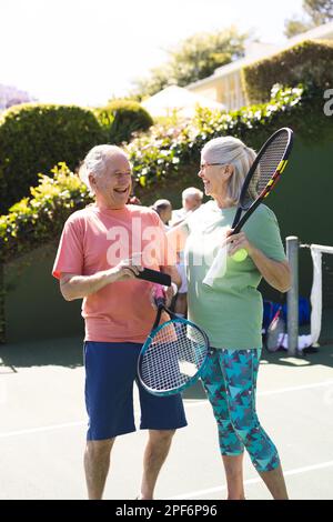 Amici anziani caucasici felici con racchette da tennis utilizzando smartphone al campo da tennis in giornata di sole Foto Stock