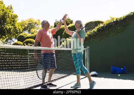 Amici anziani caucasici felici con racchette da tennis alta fiving al campo da tennis nella giornata di sole Foto Stock