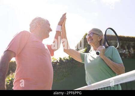 Amici anziani caucasici felici con racchette da tennis alta fiving al campo da tennis nella giornata di sole Foto Stock