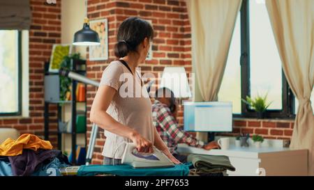 Persona stressata che lancia la blusa al socio rilassato, essendo frustrato mentre ferra i vestiti nel paese. Donna infelice che è depressa e che cerca di pulire l'appartamento, facendo la pulizia di primavera. Foto Stock