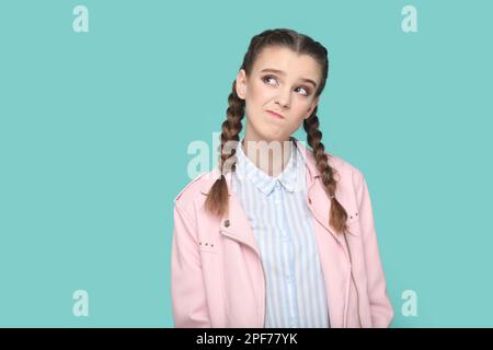 Ritratto di giovane adolescente pensosa e penosa con trecce che indossano giacca rosa guardando lontano con l'espressione confusa e perplessa. Studio in interni isolato su sfondo verde. Foto Stock