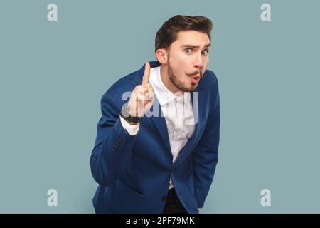 Ritratto di un astuto astuto astuto uomo con baffi in piedi con il dito sollevato, che ha un'ottima idea, indossando camicia bianca e giacca. Studio in interni isolato su sfondo azzurro. Foto Stock