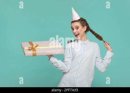 Ritratto di estremamente felice sorridente ragazza soddisfatta adolescente con trecce in camicia a righe e cono partito, tenendo presente scatola, essendo in umore festivo. Studio in interni isolato su sfondo verde. Foto Stock