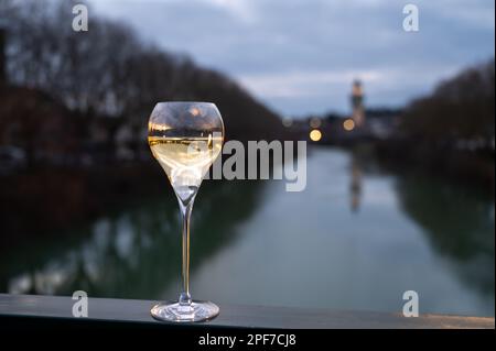 Bicchiere di vino bianco frizzante francese con champagne bolle all'aperto con vista sulle acque verdi del fiume Marne e le luci del viale Promenade in mor Foto Stock