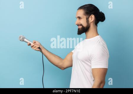 Vista laterale dell'uomo con barba che indossa una T-shirt bianca che tiene il microfono, che fa un'intervista e chiede opinioni, discutendo argomenti importanti. Studio in interni isolato su sfondo blu. Foto Stock