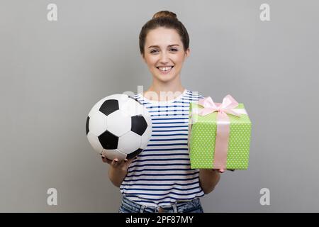 Ritratto di donna che indossa T-shirt a righe in piedi guardando sorridente alla fotocamera, tenendo fuori palla da calcio e scatola verde presente. Studio in interni isolato su sfondo grigio. Foto Stock