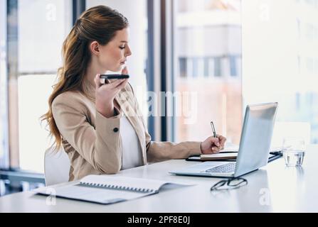 Prendere appunti durante la chiamata. una giovane donna d'affari che scrive appunti mentre usa un cellulare in un ufficio. Foto Stock