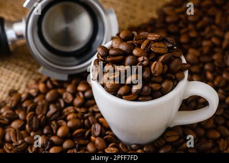 Tazza piena di chicchi di caffè interi tostati e porta-carruba sullo sfondo di un tavolo di legno, colori naturali neutri Foto Stock