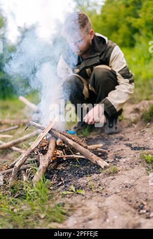 Colpo verticale di un uomo sopravvissuto in un impermeabile che inizia il fuoco utilizzando un accendino a gas sulla riva del fiume per cucinare e riscaldare la sera prima del tramonto. Foto Stock