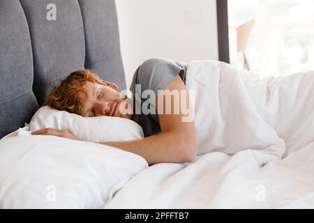Ritratto di giovane bell'uomo dormiente rosso in t-shirt grigia a letto a casa Foto Stock