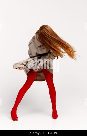 Emozioni umane impersonali. Elegante ragazza con cappotto grigio e collant rosso brillante si muove su sfondo chiaro. Moda espressiva. Concetto di fotografia d'arte Foto Stock