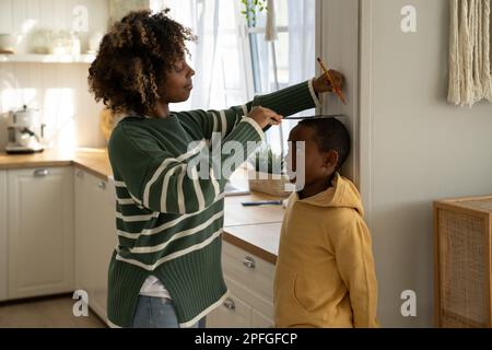 Amorevole donna nera che segue misure di aumento cambia altezza di bambino figlio sulla parete con matita a casa Foto Stock