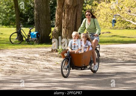 La giovane donna si sposta con la sua bici da carico con i bambini attraverso il Vondelpark di Amsterdam. Foto Stock