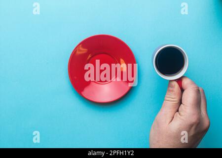 Una tazza di caffè nero da espresso tenuta da una mano su sfondo blu con un piattino Foto Stock