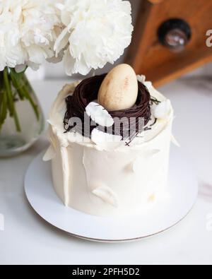 Torta di Pasqua decorata con nido d'uccello al cioccolato, uova e piume su sfondo romantico con peonie bianche e stella di legno retrò Foto Stock