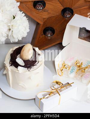 Torta di Pasqua decorata con nido d'uccello al cioccolato, uova e piume su sfondo romantico con peonie bianche e stella di legno retrò Foto Stock