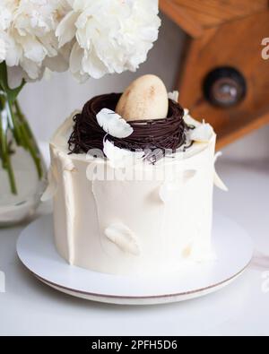 Torta di Pasqua decorata con nido d'uccello al cioccolato, uova e piume su sfondo romantico con peonie bianche e stella di legno retrò Foto Stock
