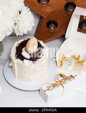 Torta di Pasqua decorata con nido d'uccello al cioccolato, uova e piume su sfondo romantico con peonie bianche e stella di legno retrò Foto Stock