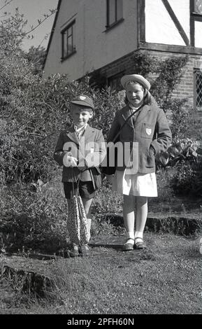 1964, storico, due bambini piccoli, fratello e sorella nella loro divisa scolastica in piedi per la loro foto, Inghilterra, Regno Unito. Tutti i classici 60s articoli scolastici nella foto, tra cui borse a mano in pelle, berretto, cappello, blazer in lana, e sandali in pelle, Foto Stock