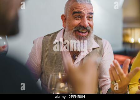 Uomo espressivo nel suo 50s, con barba lunga e braccia tatuate, gesticulando e conversando con amici sfocati. Indossa una camicia bianca e un gilet. Impostazione Foto Stock